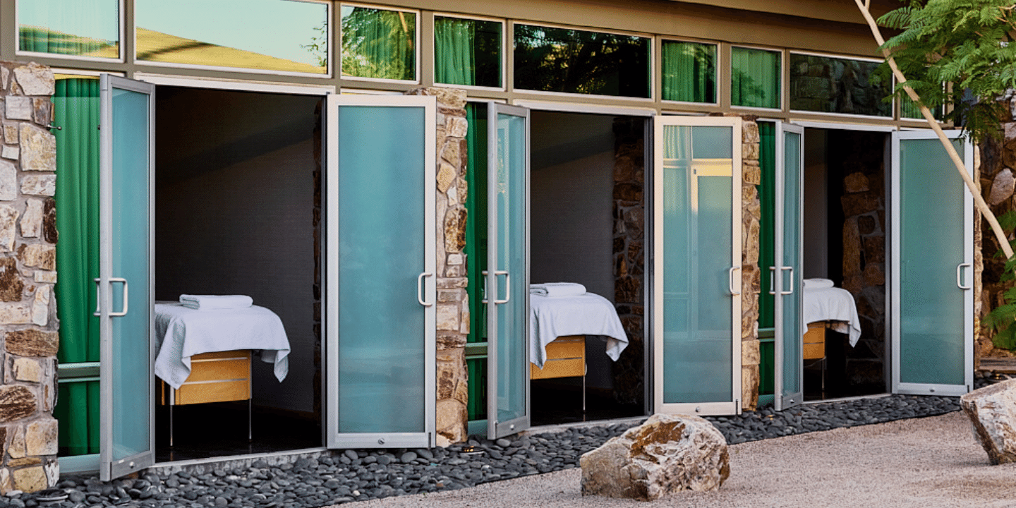 A row of frosted teal doors in a stone wall opens onto small rooms with massage tables, river stones and a boulder.