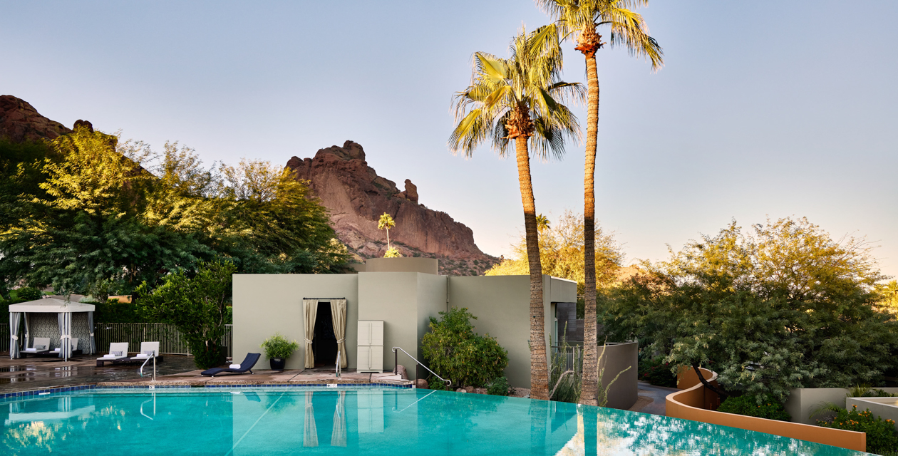 Poolside view with mountains in the background, palm trees, and a modern building surrounded by greenery under a clear sky.