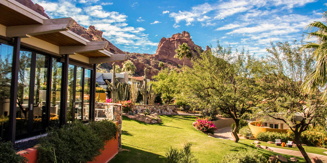 Lush garden setting with trees, blooming flowers, and a distant mountain under a bright blue sky. Modern building with large windows on the left.