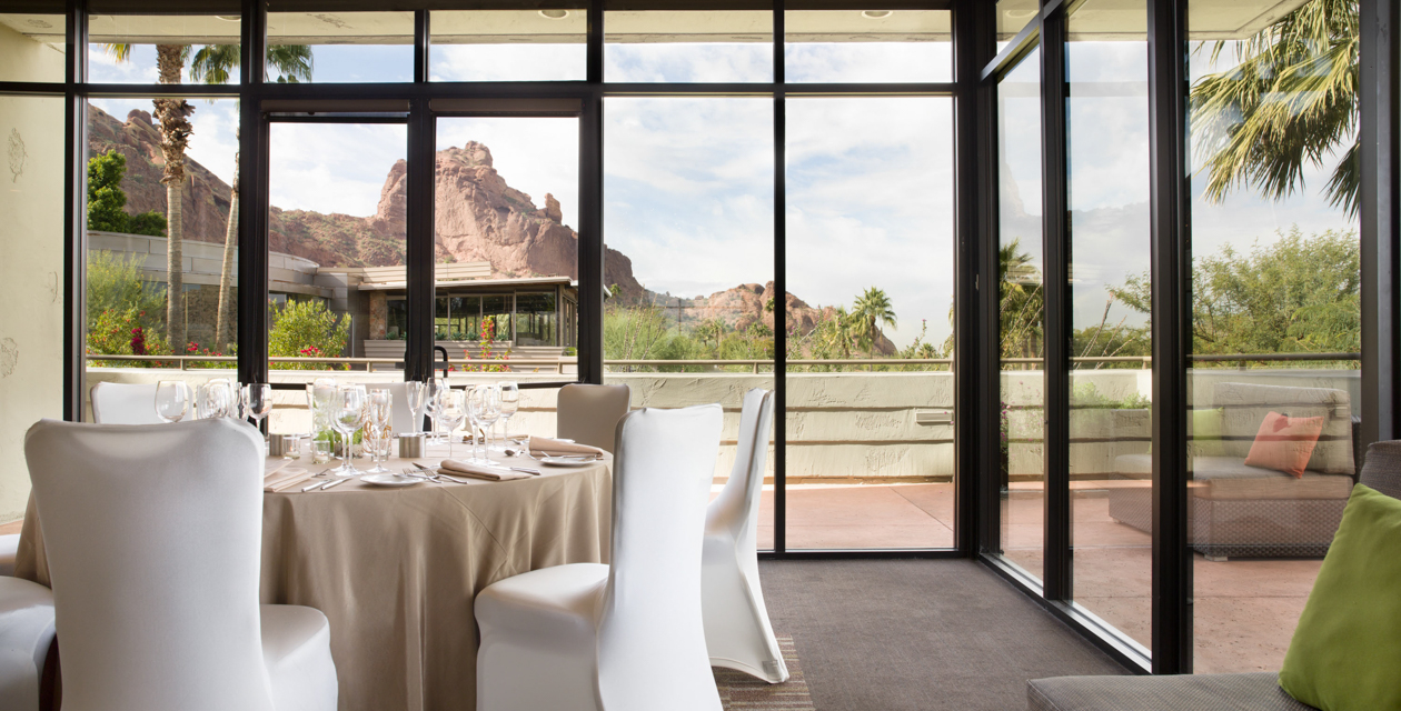 Sunny dining area with a round table set for a meal, large windows overlook a patio and scenic desert mountains in the background.