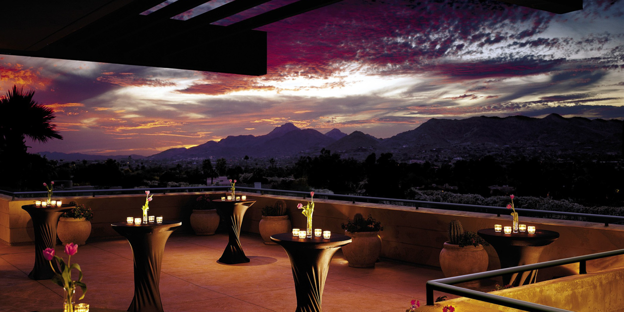 Outdoor terrace at sunset with tables and candles, overlooking a mountainous landscape under a colorful sky.