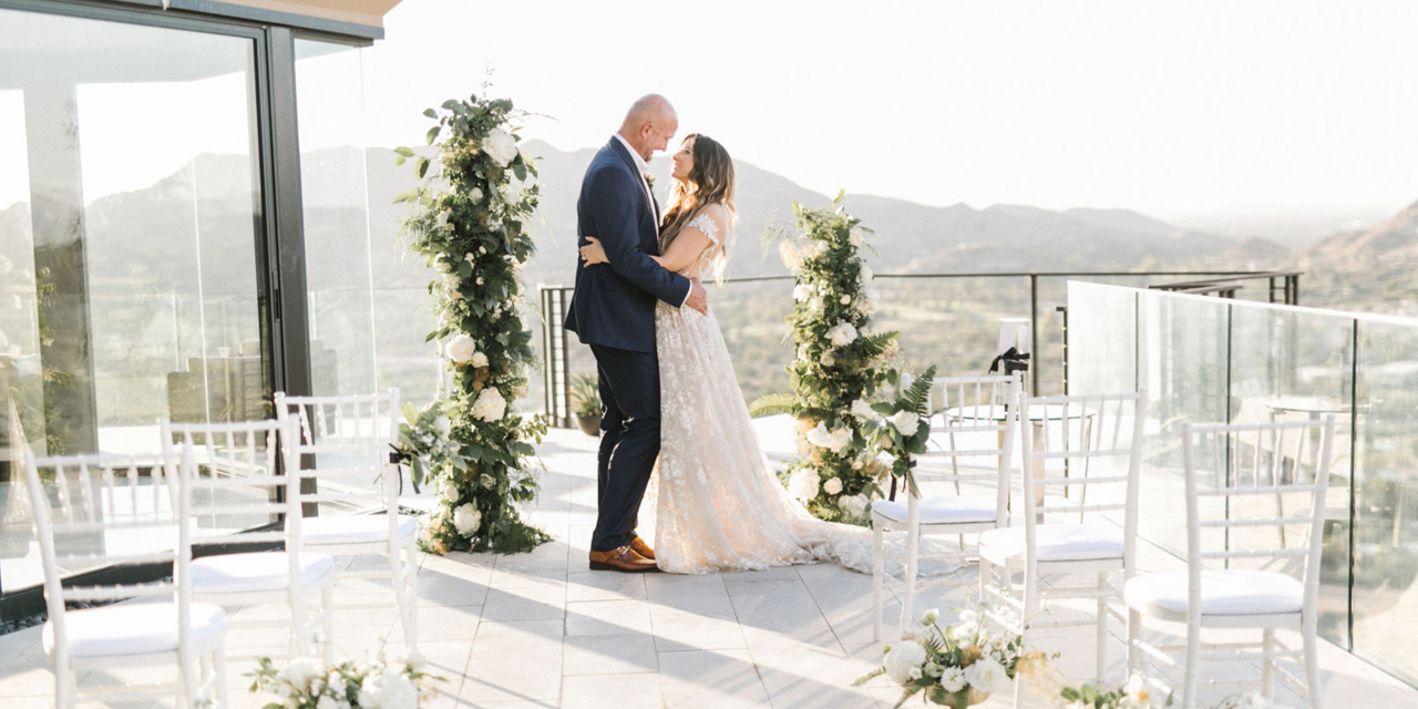 A couple embraces on a sunny outdoor wedding venue, surrounded by greenery, with mountains in the background.