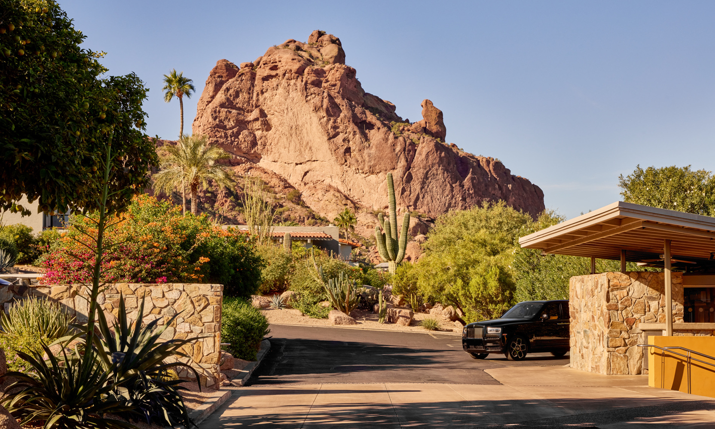 A road curves past a stone-walled covered entrance with a timber canopy and a black SUV, with shrubs, cacti and a rock peak rising behind.