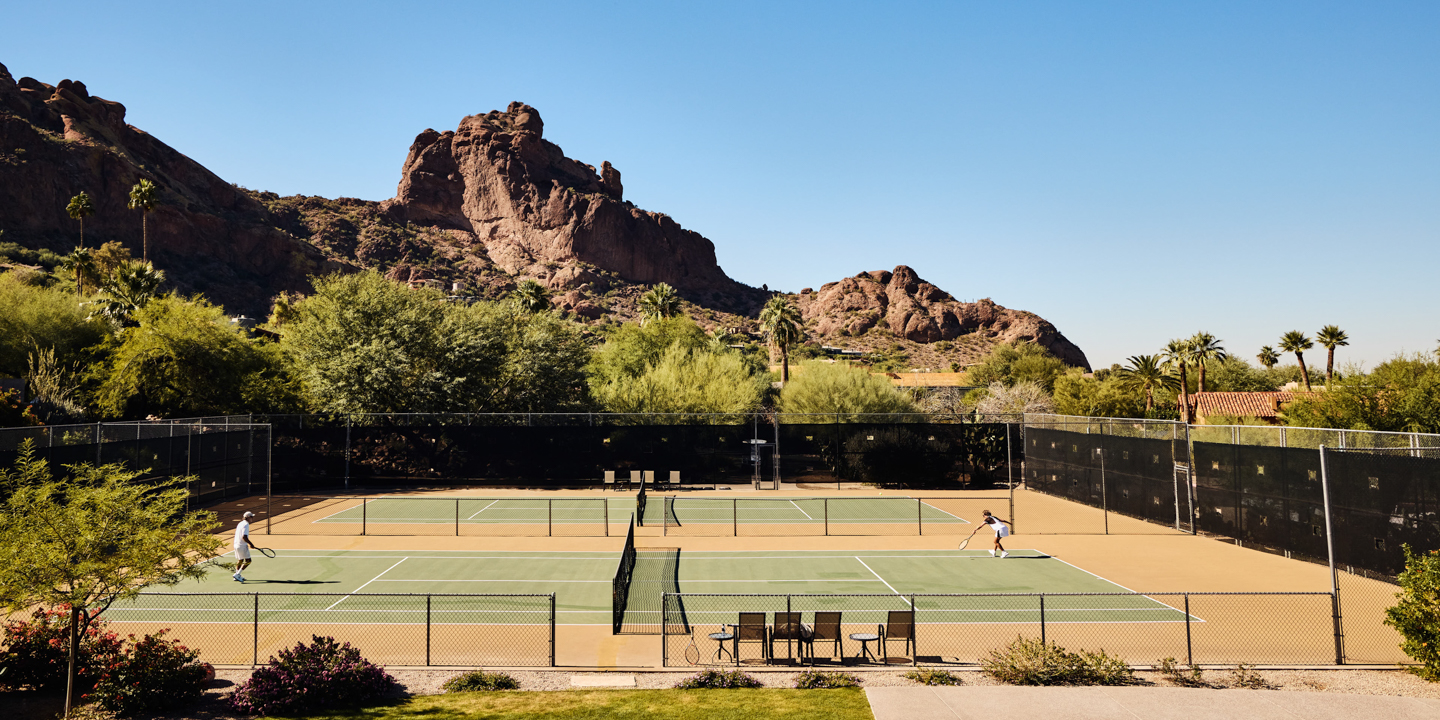 Tennis courts with green surfaces and tan surrounds sit behind black windscreens, with two players mid-game and a rust-red ridge beyond.