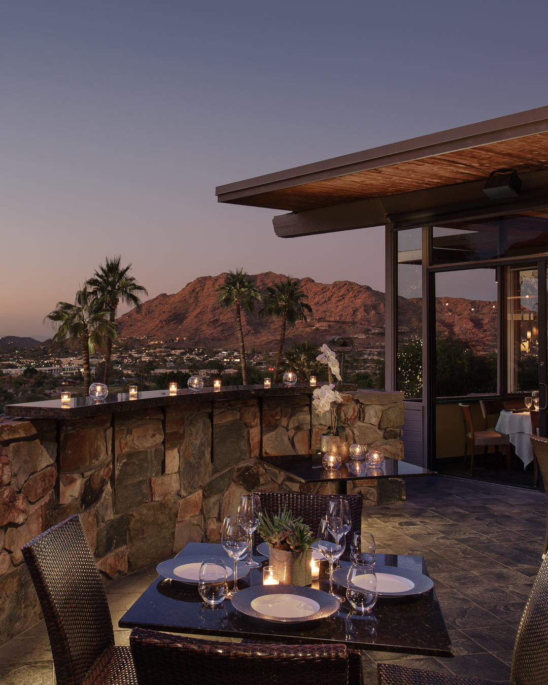 Rooftop restaurant at sunset with lit candles on tables, overlooking mountains and palm trees in the distance.