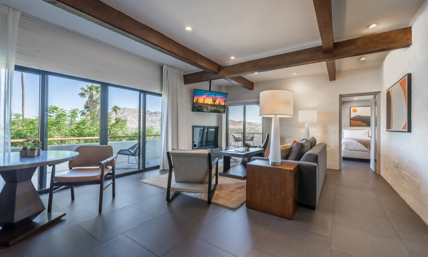 A grey sofa, leather armchairs and a round table sit under exposed timber beams, with sliding glass doors opening to a balcony with a mountain view.