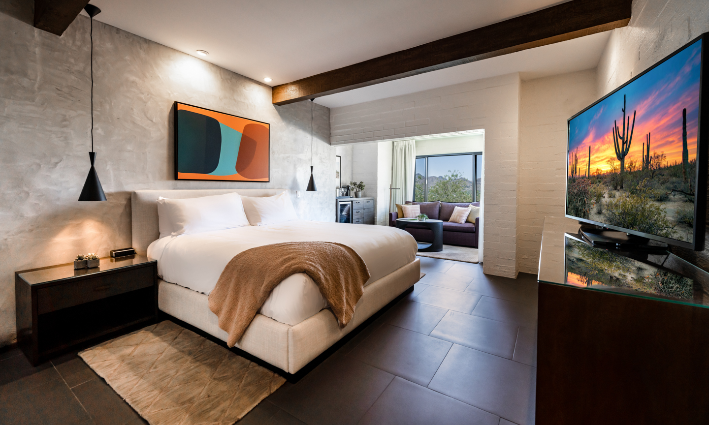 A bed with white linens and a tan throw sits on dark tile below black pendant lights and orange and teal artwork.