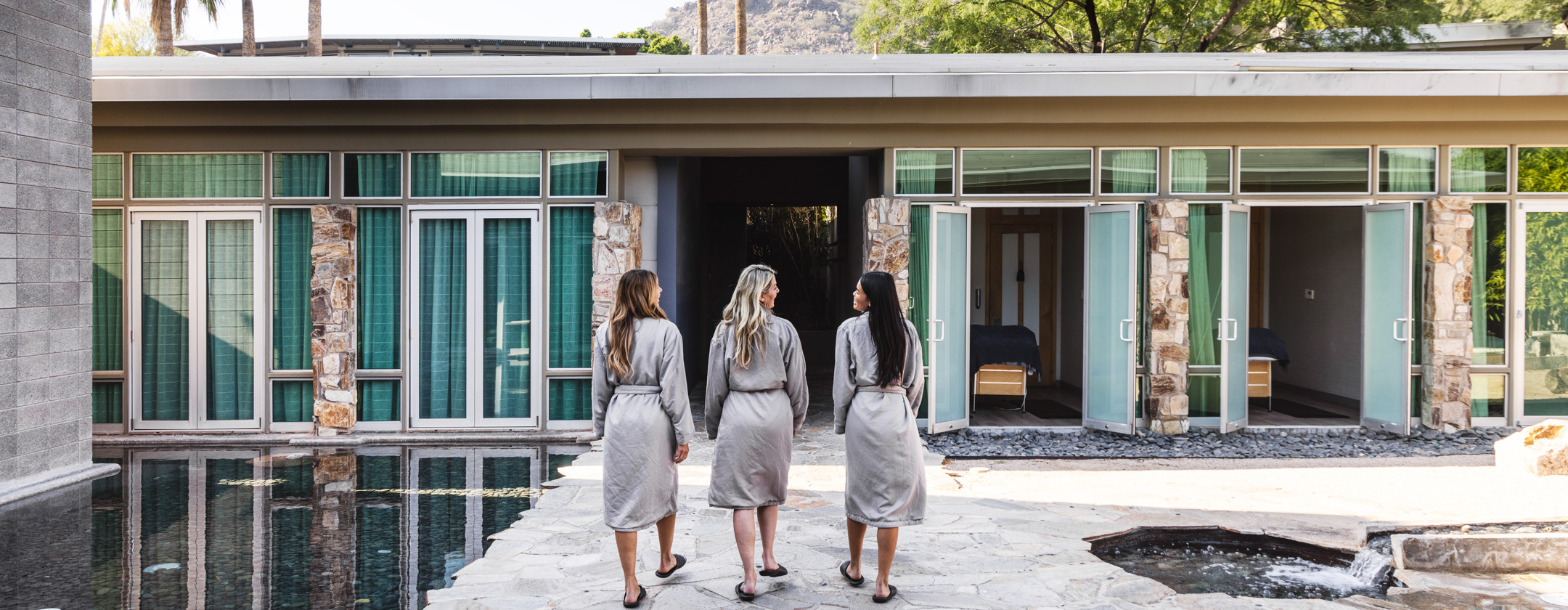 Three people in grey robes and sandals walk across stone paving toward a flat-roofed building with green curtains, doors and treatment tables.