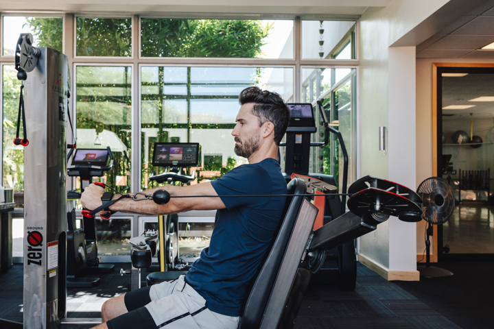 A man sits at a cable chest-press machine in a gym with cardio machines and screens, tall windows showing greenery behind.