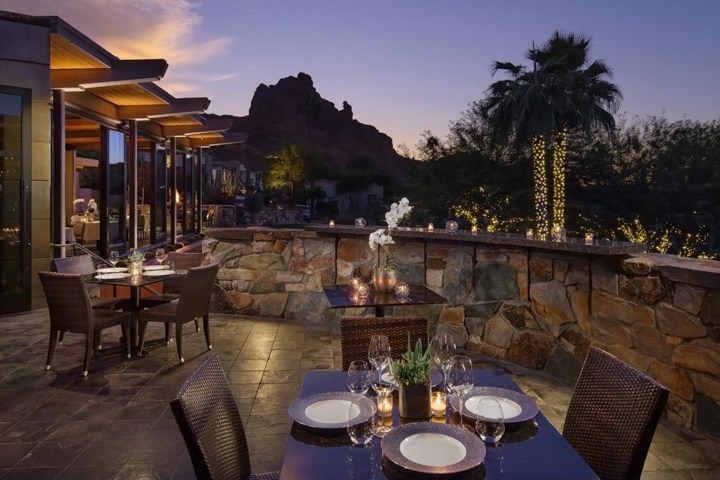 A dusk terrace with tables, plates, glasses, candles and white flowers sits behind a stone wall with string lights and mountain silhouette.
