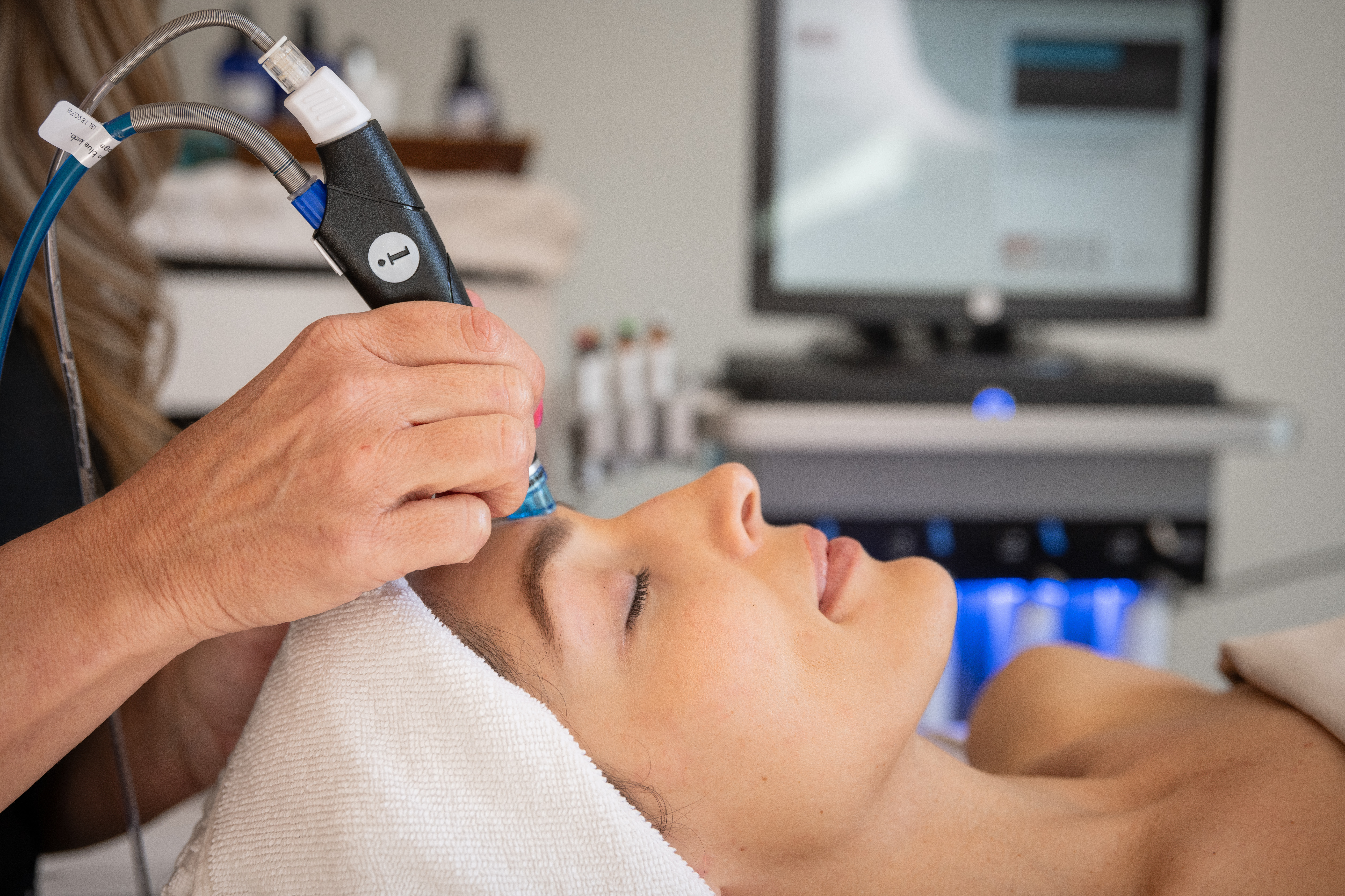 A white towel wraps the head as a hand holds a device against the forehead, with a machine and blue light behind.