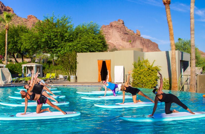 People practicing yoga on paddleboards in a pool, with mountains and palm trees in the background.
