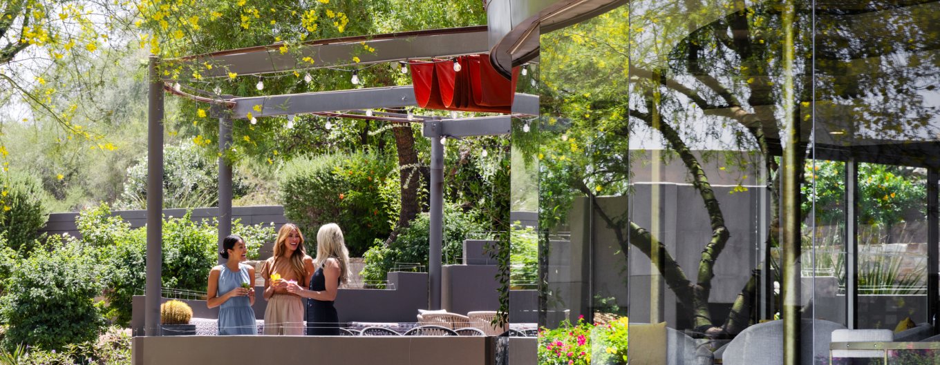 Three girlfriends enjoying craft cocktails and a laugh on jade bar patio surrounded by lush desert landscape.