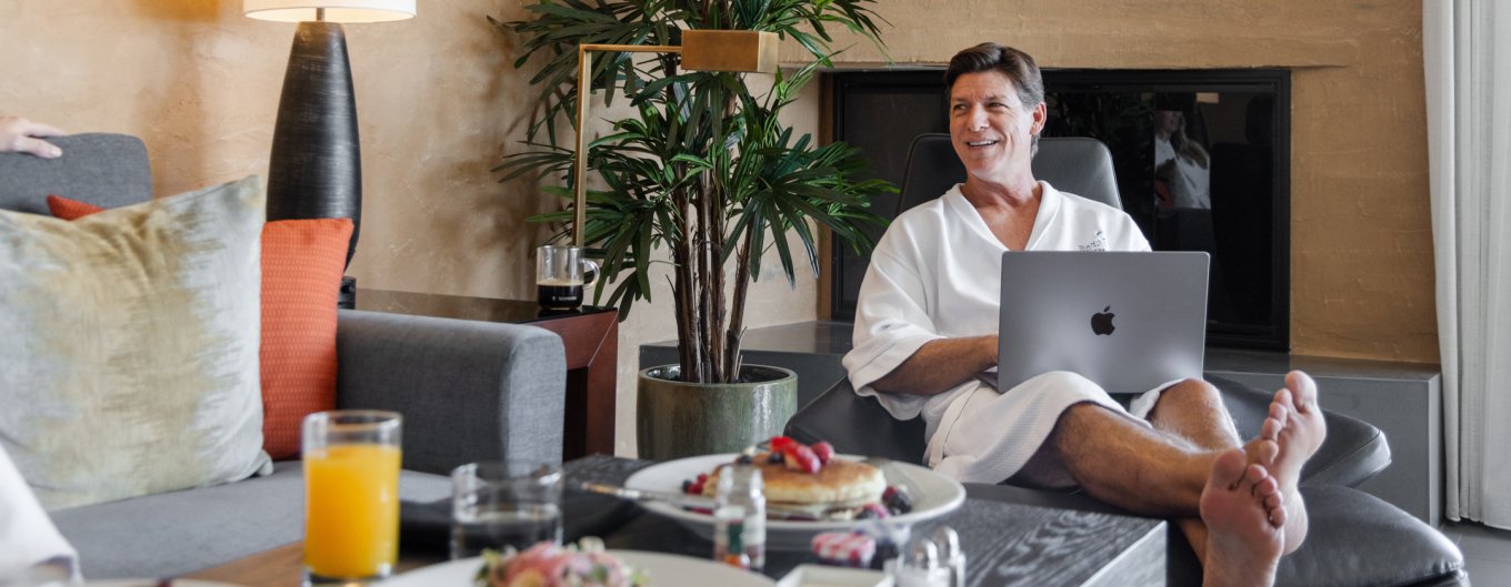 Man in robes sitting by in-room breakfast and using an apple computer.