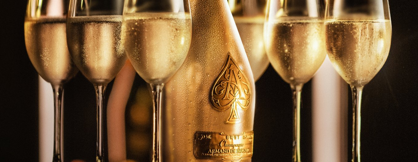 Gold bottle of Armand de Brignac Champagne against black background surrounded by filled glasses of champagne.