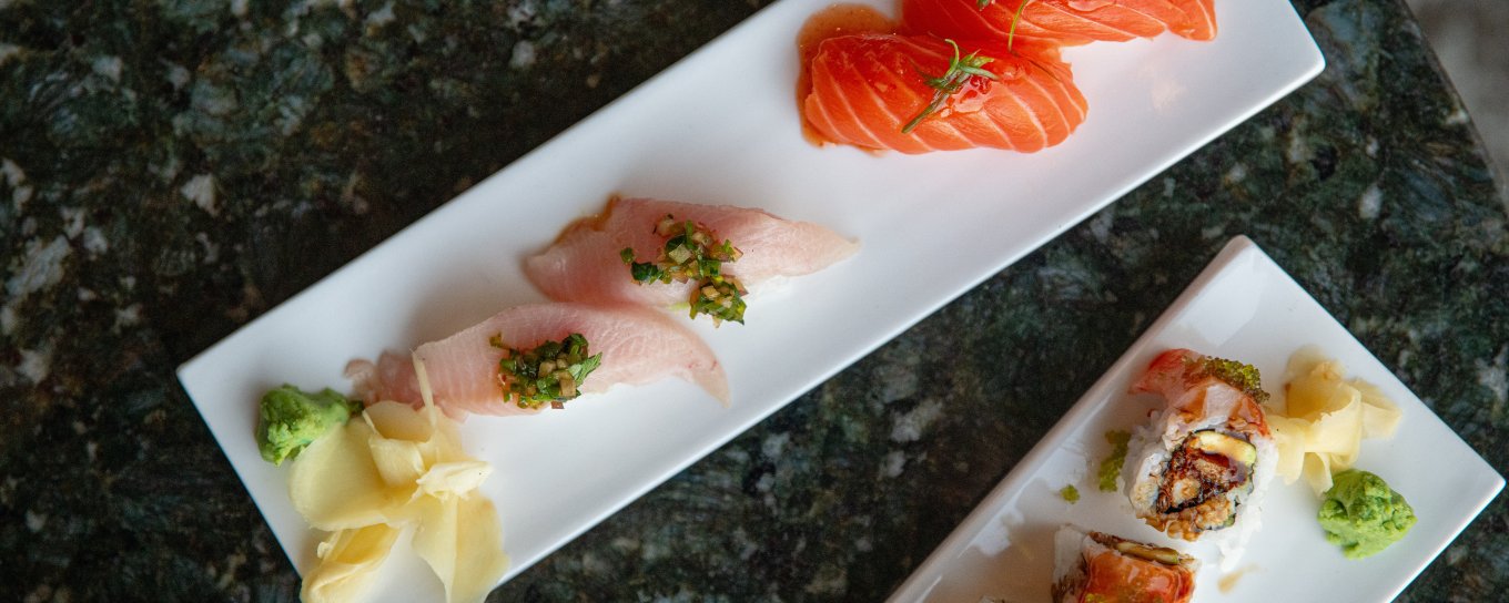 Three rectangular plates of sushi with salmon, white fish, and rolls, garnished with wasabi and ginger, on a dark marble table.
