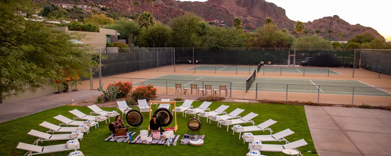 Circular arrangement of lounge chairs on grass, surrounded by desert landscape and mountains at sunset. Tennis courts in the background.