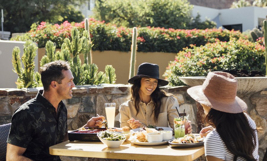 Three people sit at an outdoor patio table enjoying a meal, surrounded by plants and distant mountains under a bright sky.