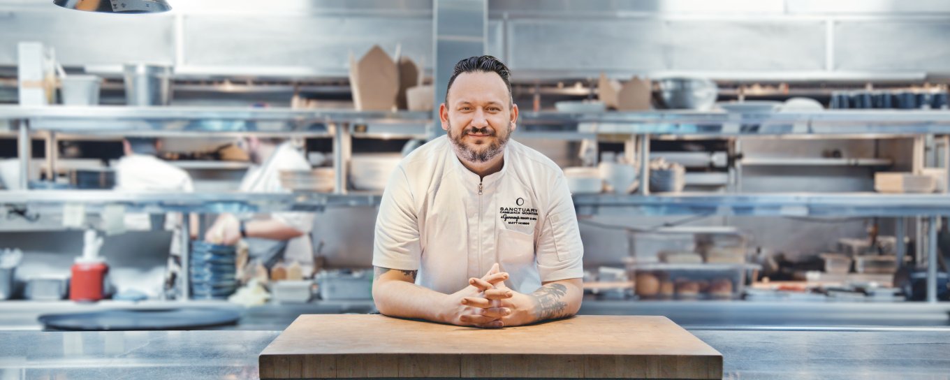 Chef in a white uniform leans on a wooden counter in a bustling, stainless steel kitchen.