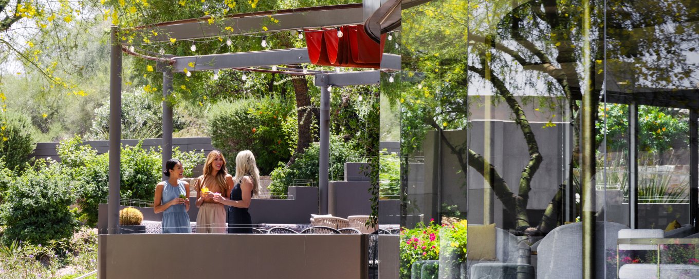 Modern outdoor café with people seated at tables, surrounded by lush greenery and string lights. Reflective glass facade curves on the right.