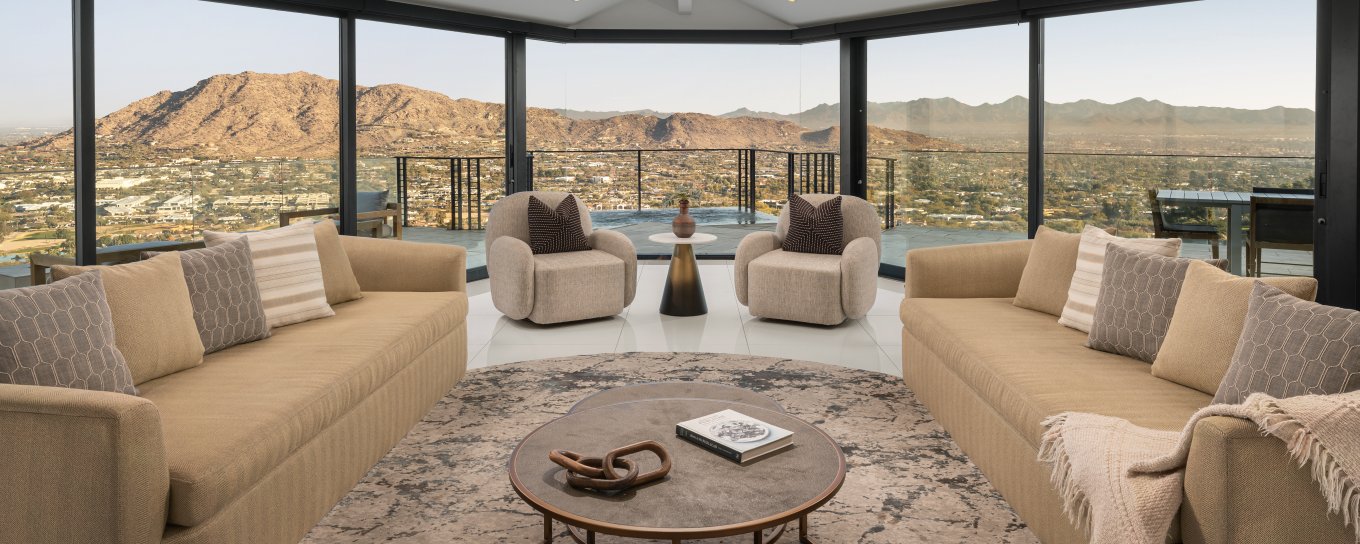 Elegant living room with large windows, mountain view, two beige sofas, two armchairs, and a round coffee table on a patterned rug.
