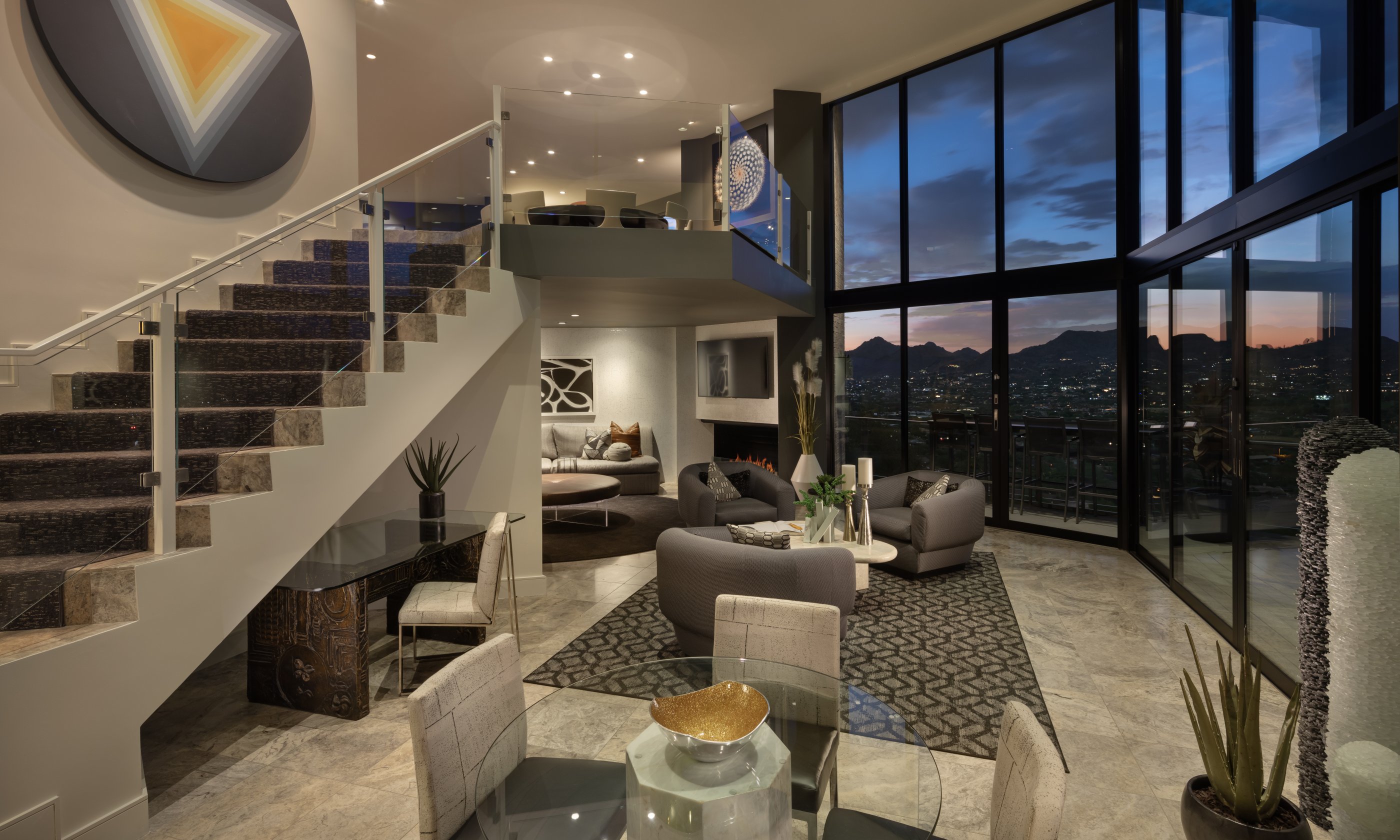 Modern penthouse interior with a glass wall showing a mountain sunset, stylish furniture, and a large geometric wall art next to the staircase.