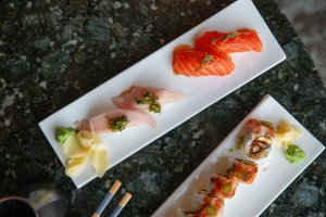 Three rectangular plates of sushi with salmon, white fish, and rolls, garnished with wasabi and ginger, on a dark marble table.