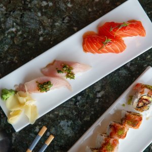 Three rectangular plates of sushi with salmon, white fish, and rolls, garnished with wasabi and ginger, on a dark marble table.