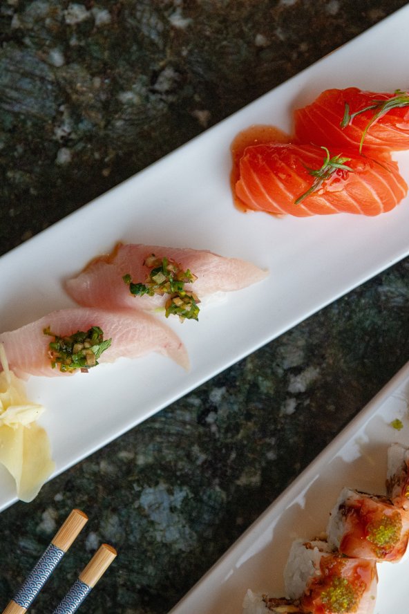 Three rectangular plates of sushi with salmon, white fish, and rolls, garnished with wasabi and ginger, on a dark marble table.