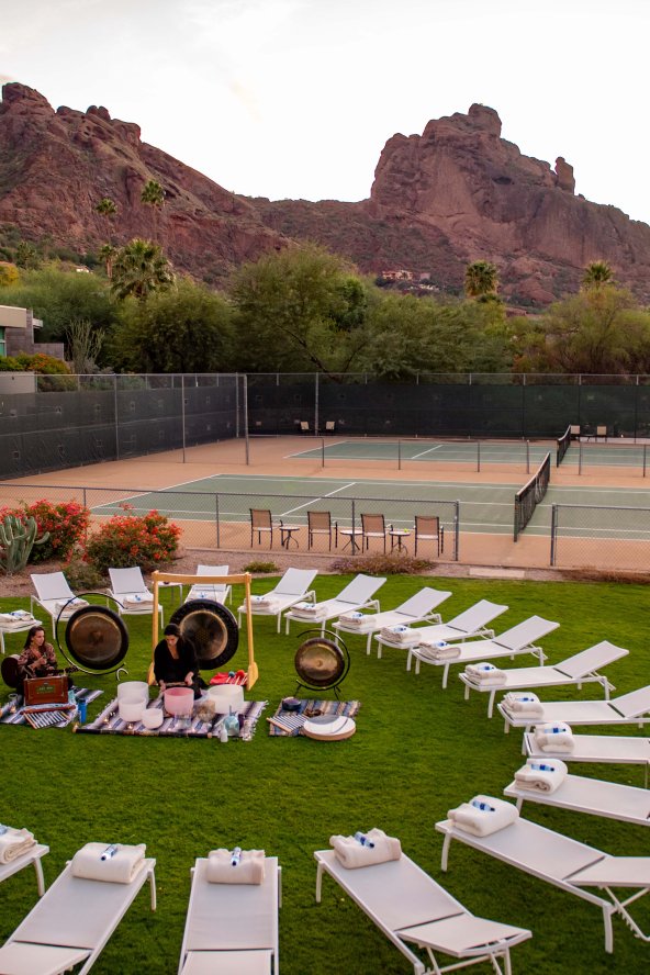 Circular arrangement of lounge chairs on grass, surrounded by desert landscape and mountains at sunset. Tennis courts in the background.