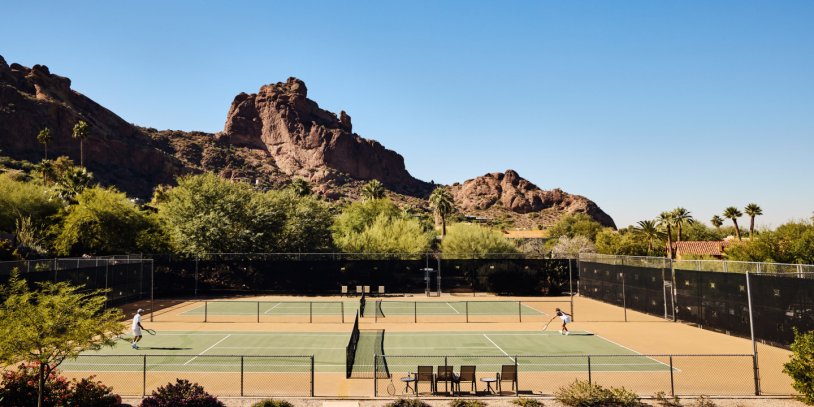 Tennis courts with players, set against desert mountains and clear blue sky.