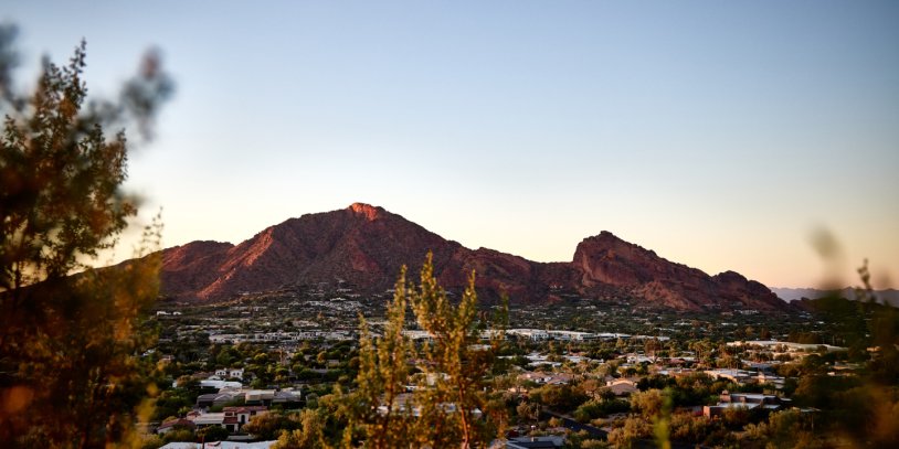 Mountain range at sunset with a clear sky, surrounded by a valley of houses and greenery, framed by tree branches in the foreground.