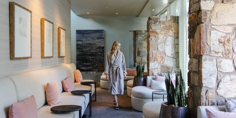 Woman in robe walking through a modern spa lounge with beige sofas, pink cushions, and stone walls.