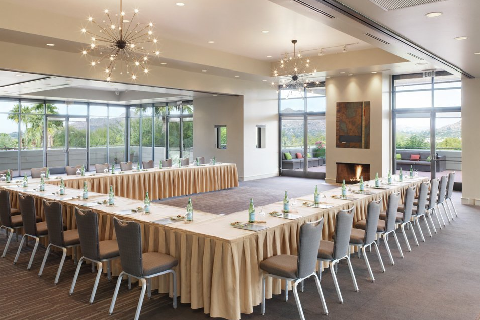 Elegant conference room with U-shaped table setup, beige tablecloths, and bottled water. Large windows, chandeliers, and scenic outdoor view.