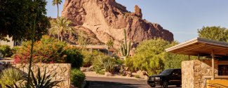Desert landscape with a prominent rocky mountain in the background, surrounded by lush greenery and a parked black car in the foreground.
