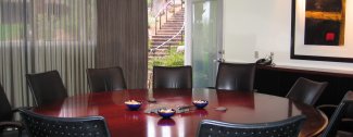A conference room with a large oval table, surrounded by black chairs. A painting and window with a view of stairs are in the background.