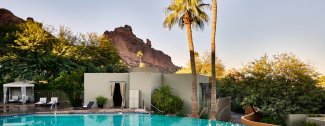 Poolside view with mountains in the background, palm trees, and a modern building surrounded by greenery under a clear sky.