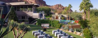 Modern resort with pool, palm trees, and mountain backdrop. White chairs on the lawn overlook the scene.