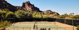 Tennis courts with players, set against a backdrop of rocky hills and greenery on a clear day.