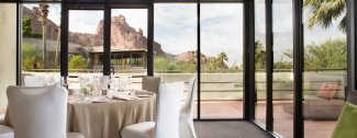 Sunny dining area with a round table set for a meal, large windows overlook a patio and scenic desert mountains in the background.