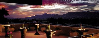 Outdoor terrace at sunset with tables and candles, overlooking a mountainous landscape under a colorful sky.
