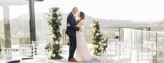 A couple embraces on a sunny outdoor wedding venue, surrounded by greenery, with mountains in the background.