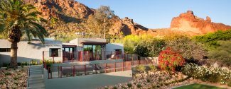 Modern house with large windows, surrounded by desert landscaping, palm trees, and a rocky mountain in the background under a clear blue sky.